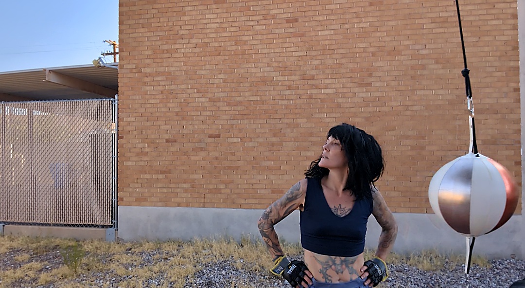 Bre Sheehan outdoors wearing boxing gloves beside a speed bag, hands on hips